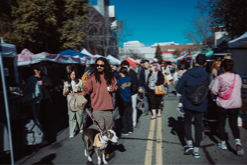 A stylish young Filipino man walks with his pet dog while visiting a busy Farmer's Market in San Francisco